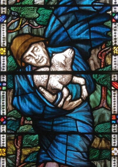 Stained glass depicting a figure in blue holding a lamb, surrounded by trees and colorful geometric patterns