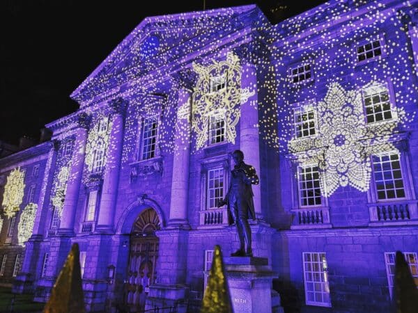 Trinity College illuminated with vibrant purple lights and intricate patterns, featuring a statue in the foreground