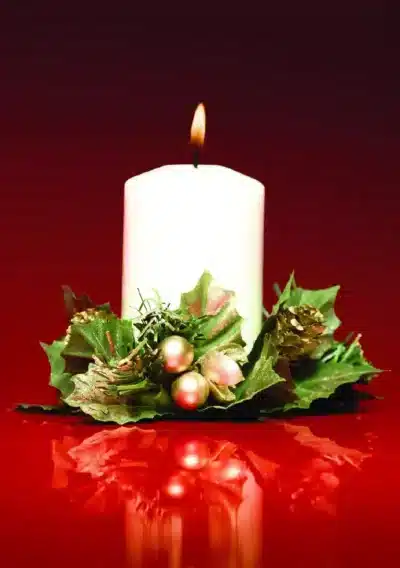 A lit white candle surrounded by green foliage and ornaments, reflecting on a red surface, evokes a festive mood for The Whit